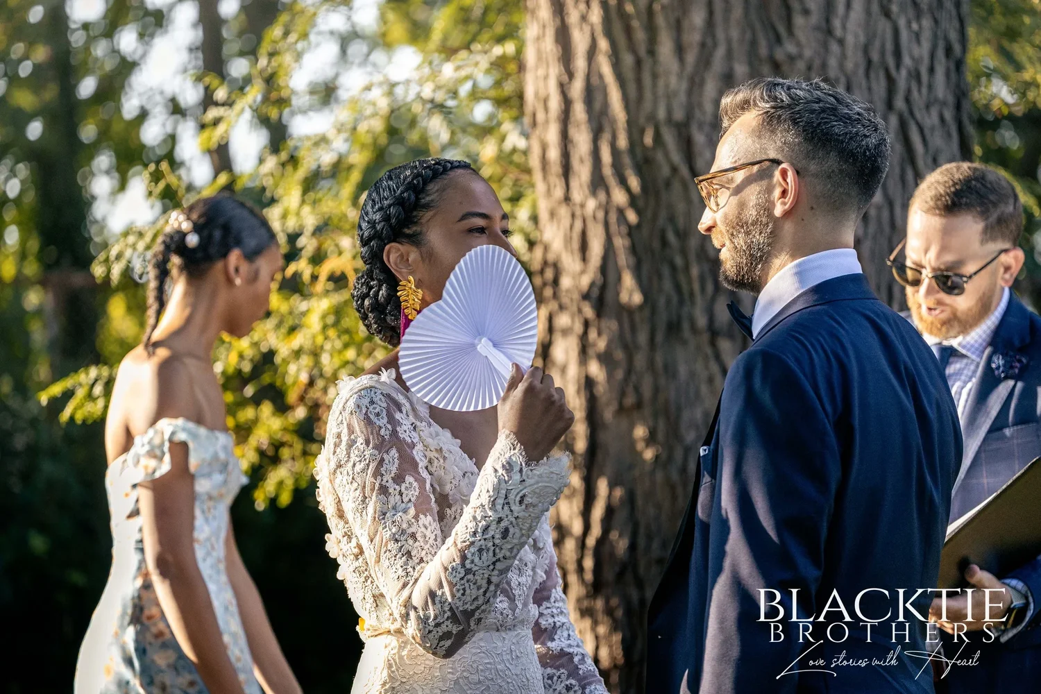 Ciara laughing behind a white paper fan during the ceremony while Drew and the officiant smile, a large tree trunk behind them