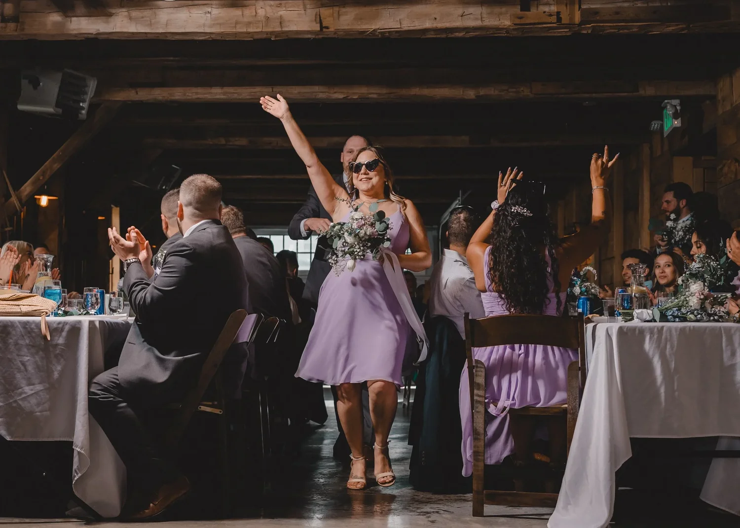 A bridesmaid in a lilac dress and sunglasses throwing her arms up during the wedding party entrance in the Grand Barn Hall
