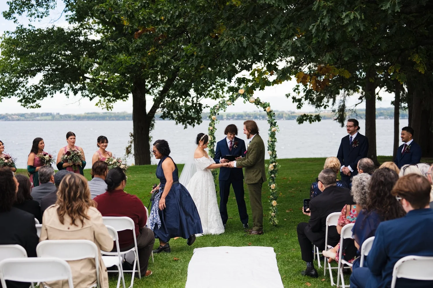 Marseille and Jason at their rose arch on the Belhurst lawn, with Seneca Lake behind them and guests on white chairs under the trees