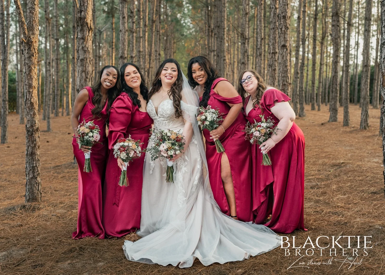 Alyssa stands smiling with members of her wedding party in burgundy dresses among the pine trees.