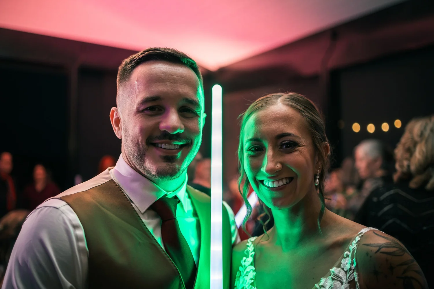 Vincent and Heather grinning in the glow of green and pink light sabers on the dance floor