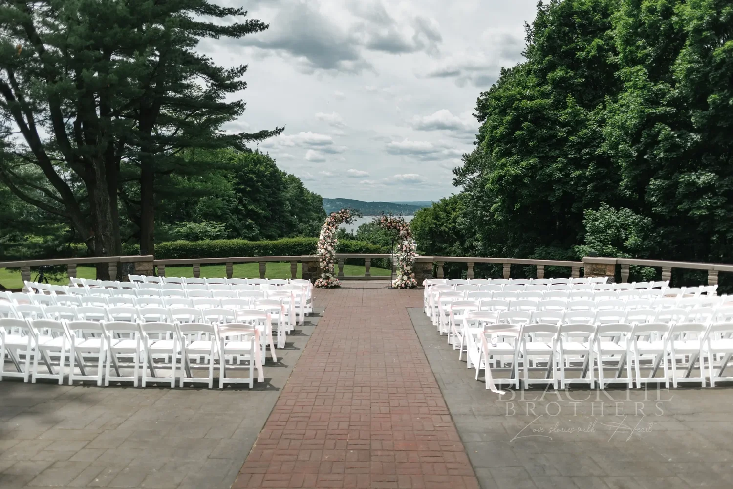 The ceremony terrace at Tappan Hill Mansion set for a wedding: rows of white chairs either side of a brick aisle leading to two floral arches at the stone balustrade, with trees and the Hudson River beyond
