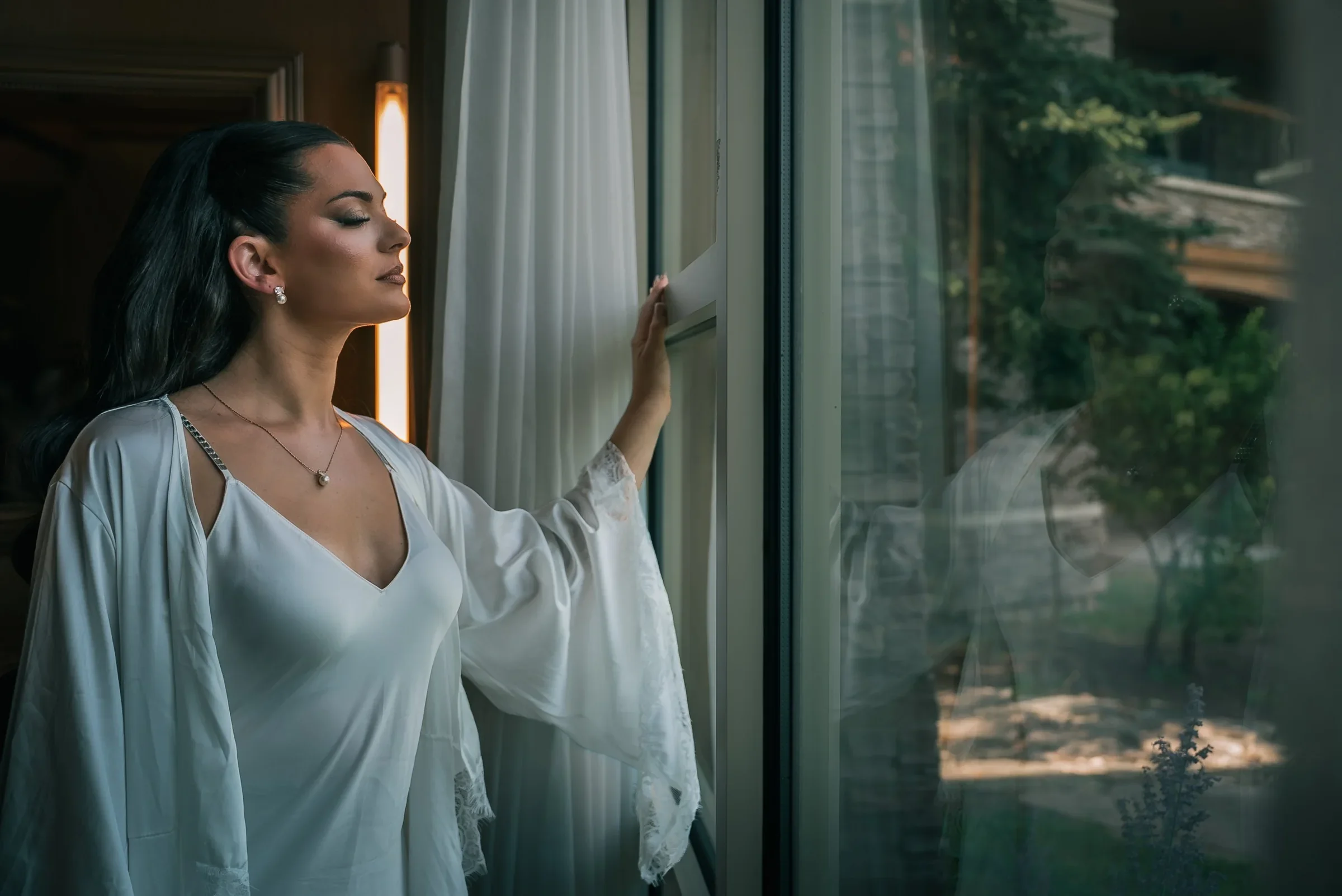 Vanessa in a white robe with lace sleeves, eyes closed in the window light, her reflection in the glass