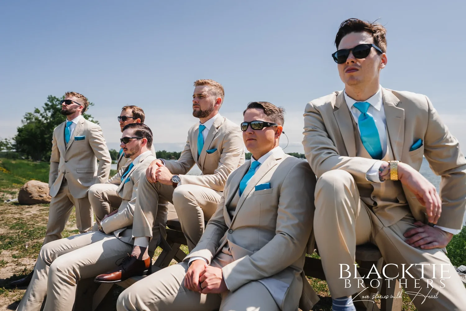 The groomsmen in tan suits and teal ties sitting on and around a picnic table at the shoreline, several in sunglasses, the lake behind them