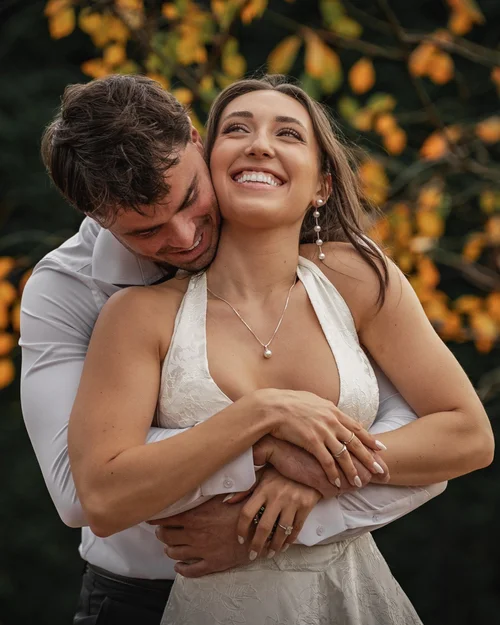 A smiling bride leans into her partner as they embrace in front of golden autumn leaves.