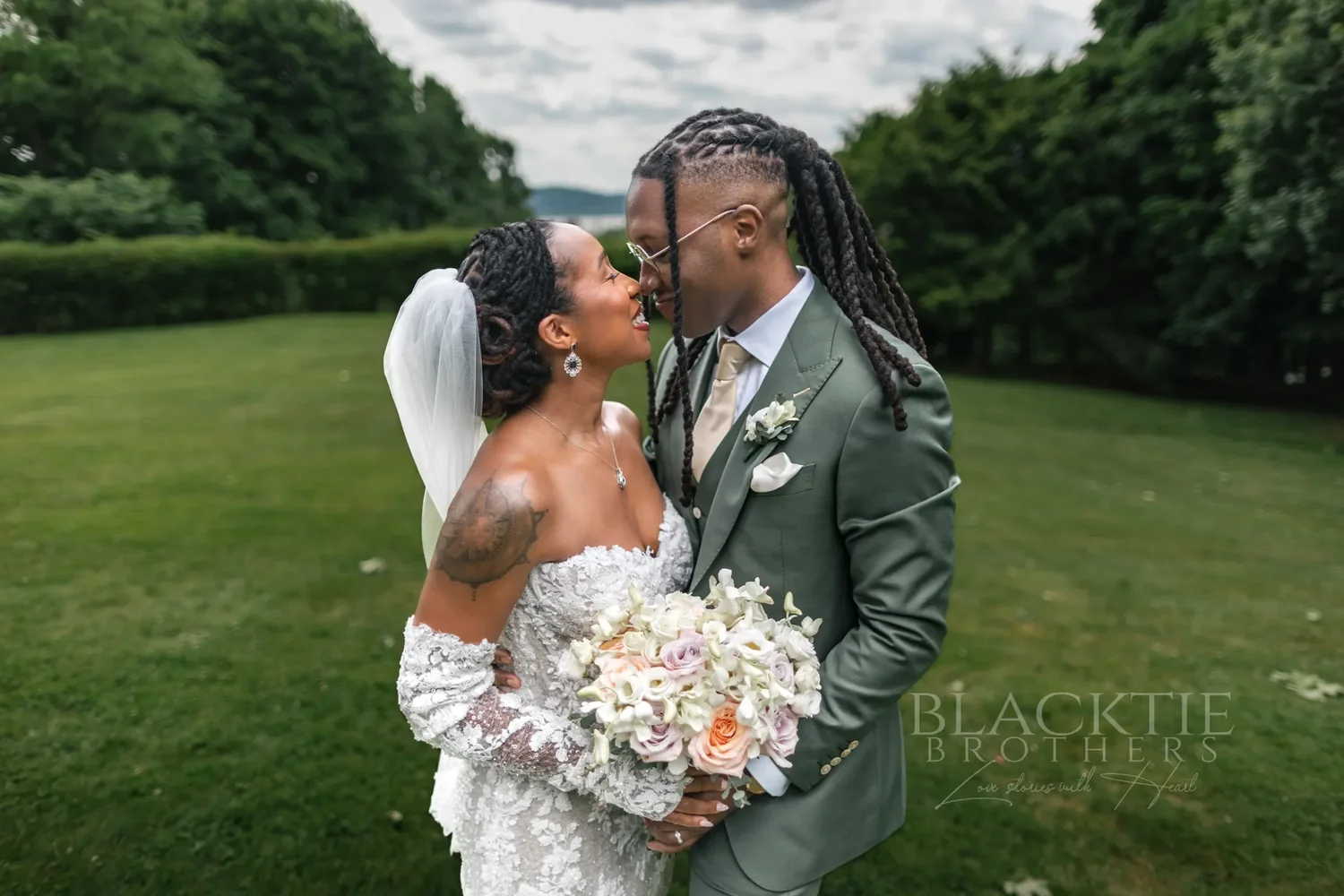 The couple kissing on the lawn, the bride holding her bouquet, with hedges and trees behind them
