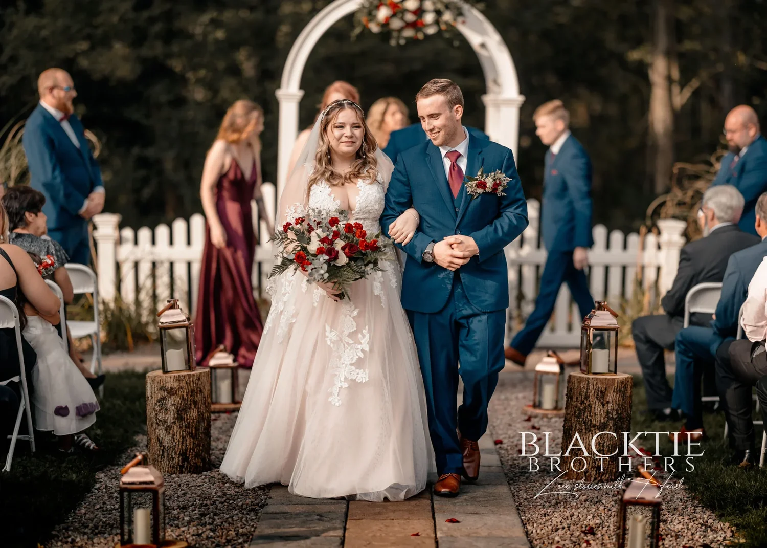 Jennifer and Jared walking back down the aisle arm in arm after the ceremony, past copper lanterns on log rounds, guests seated on either side and the white arbor behind them