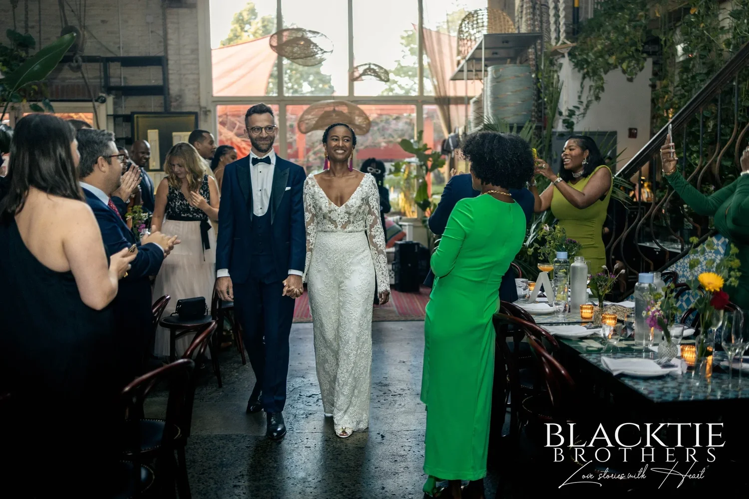 Ciara and Drew walking into the main dining room at Fandi Mata as guests stand and cheer at long candlelit tables, big windows glowing at the far end