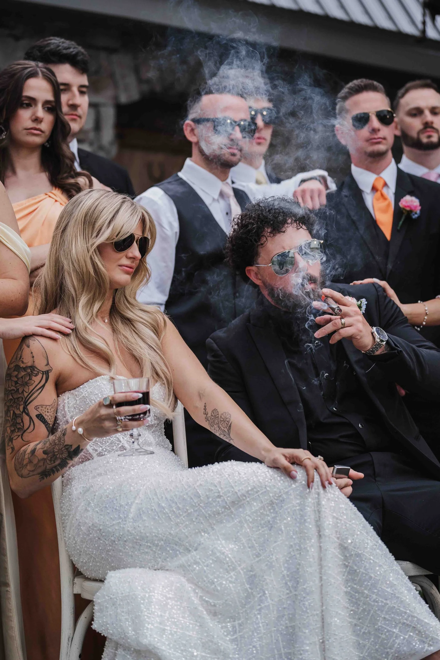 Kari and Josh sit in sunglasses in front of their wedding party, Kari holding a glass of red wine while Josh smokes a cigar.