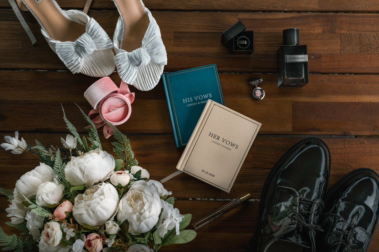 Two vow books, wedding shoes, a cologne bottle and a bouquet of white flowers laid out on a wooden floor
