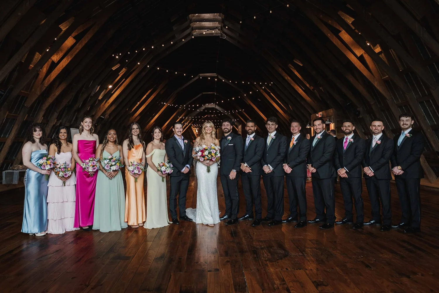 Kari, Josh and their wedding party pose in a line inside the barn under string lights.