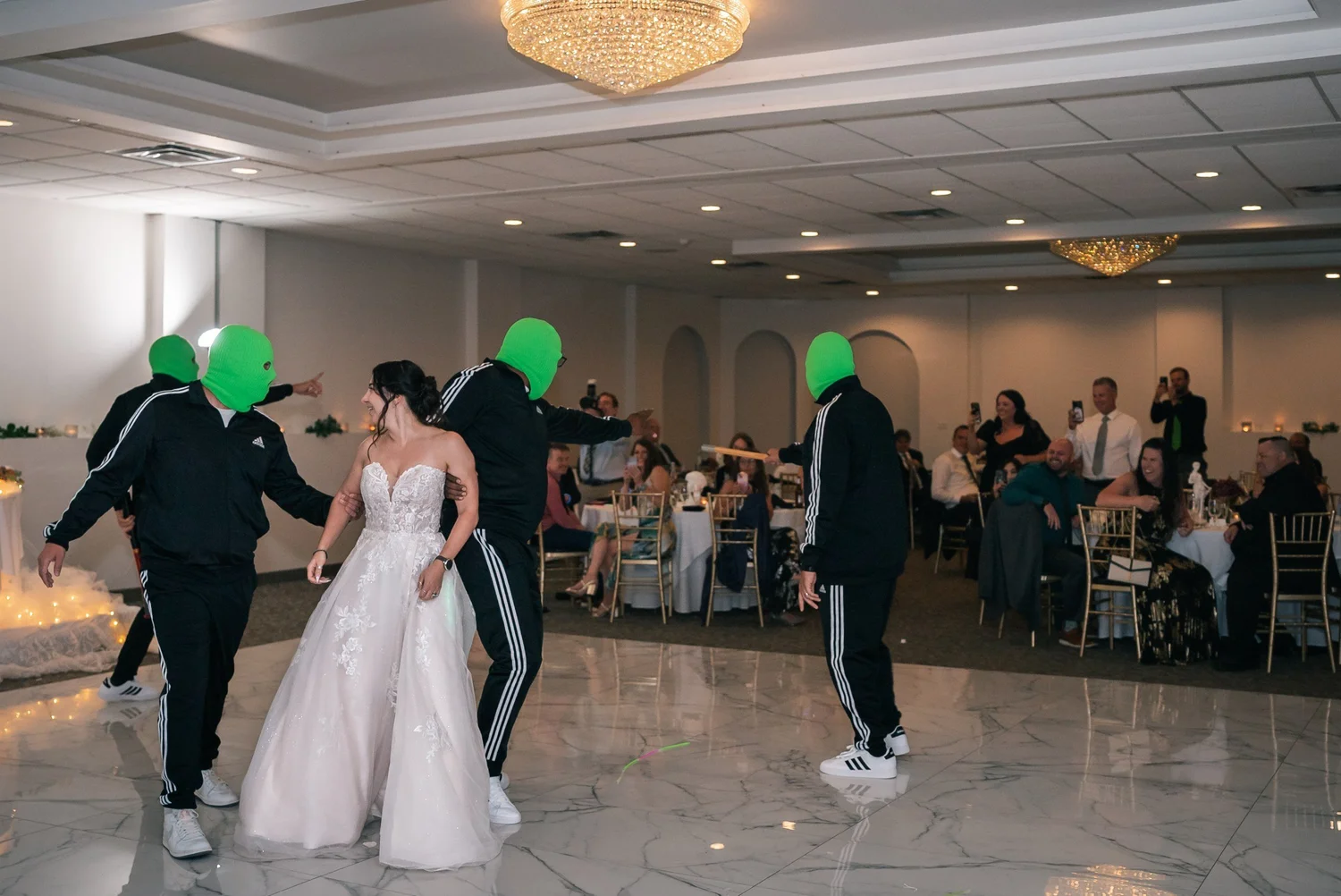 Laura dancing on the ballroom floor with a crew of guests in green full-face masks and black tracksuits
