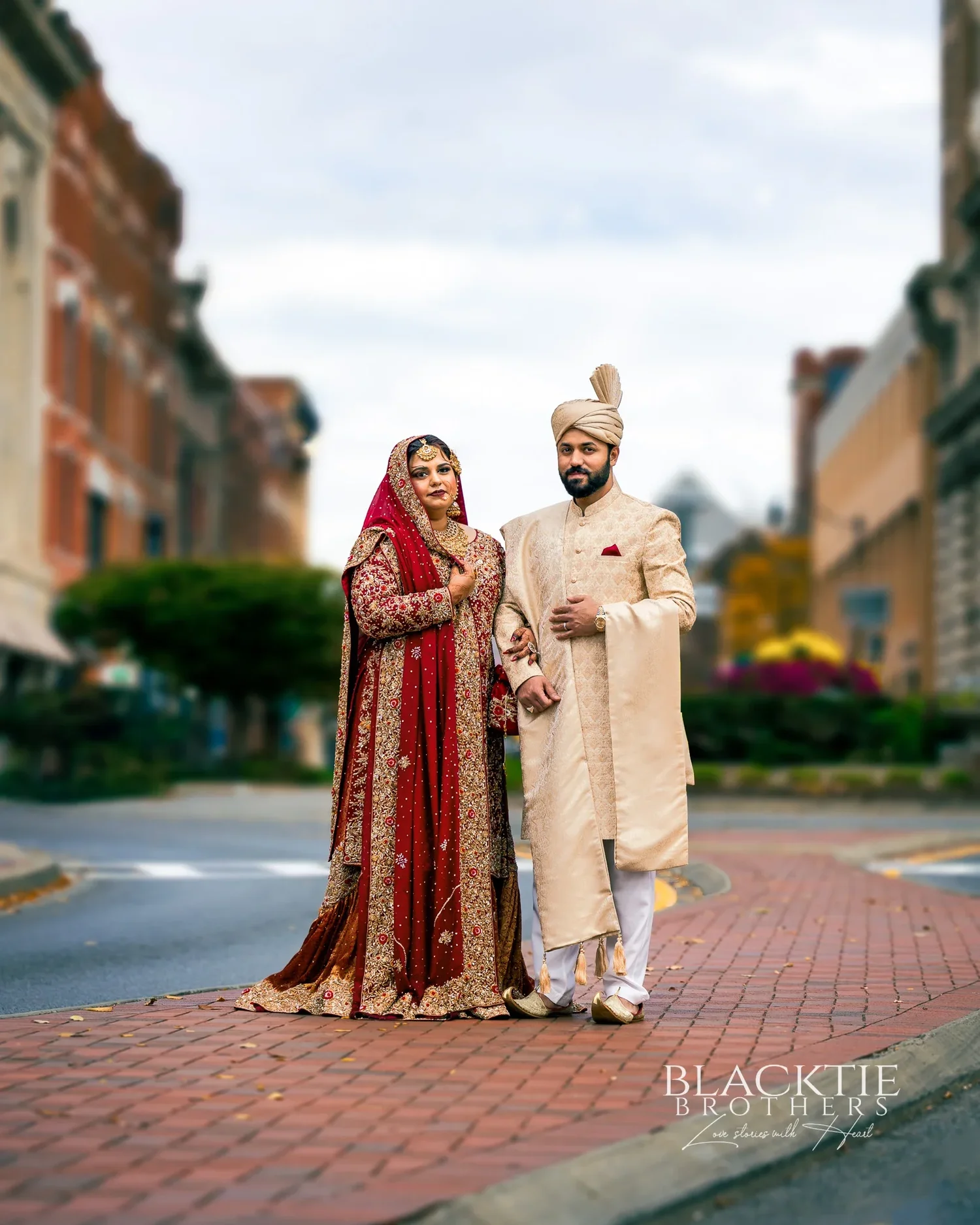 A bride in a red and gold embroidered outfit and a groom in an ivory sherwani and turban standing together on a brick crosswalk on a downtown street lined with brick buildings