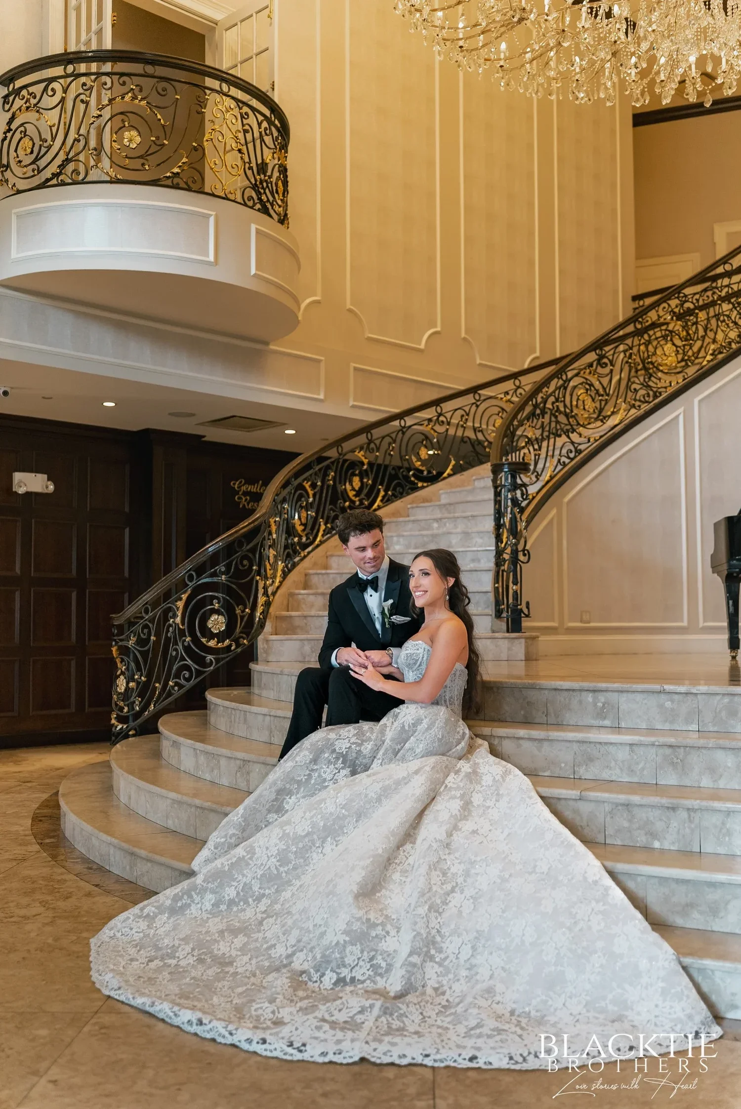 Leah and Nicholas sitting together on the marble staircase, her lace gown spread down the steps, the curved balcony and wrought-iron railings above