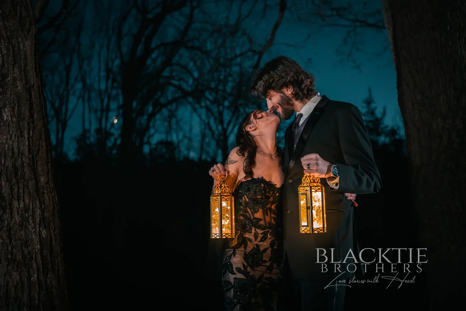 Maddy in a black lace wedding gown and Dan kissing in the woods at blue hour, both holding glowing lanterns, at Stratton Creek Inn
