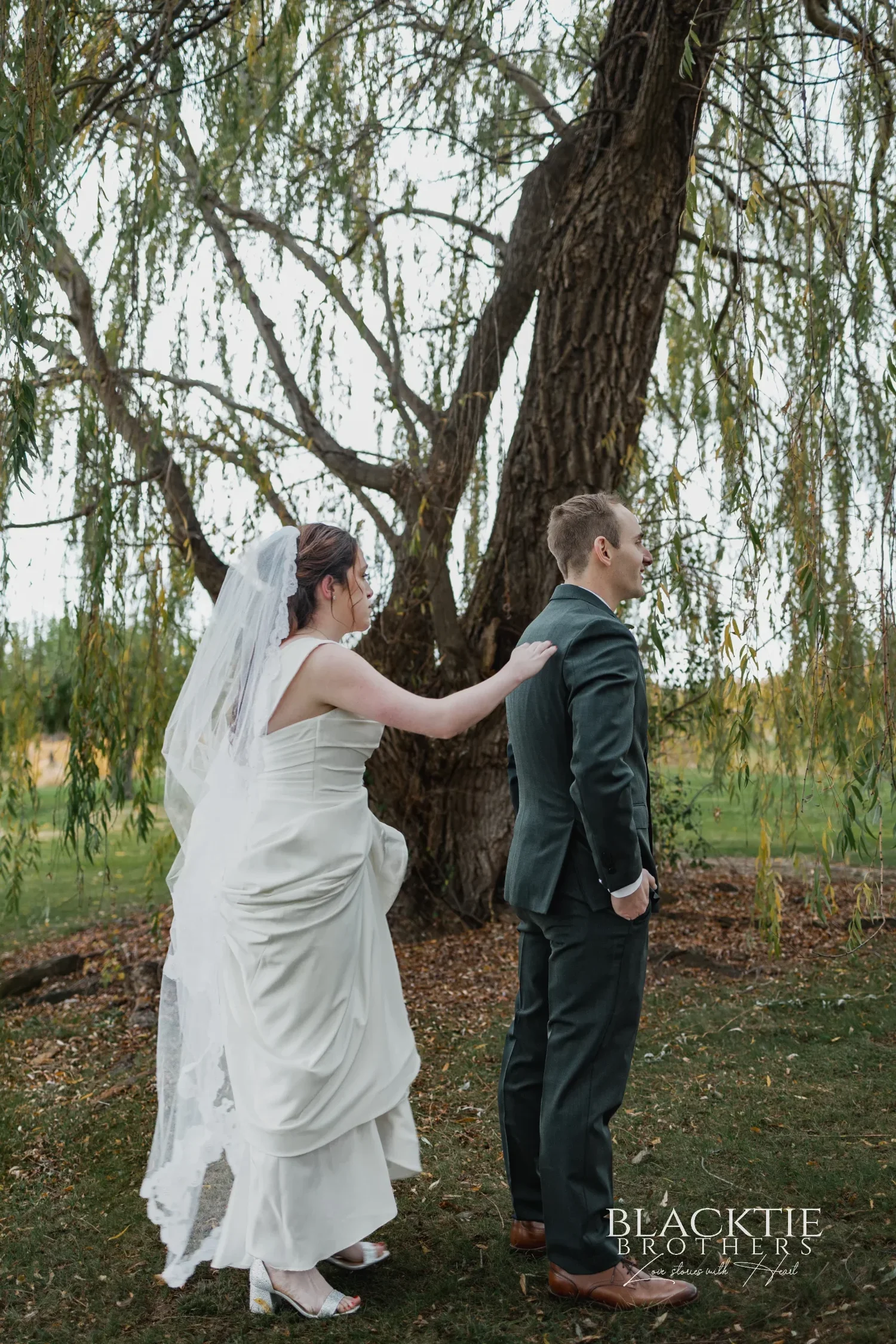 Anya and Gabe standing back to back beneath a willow tree at Glenora for a first touch before the ceremony