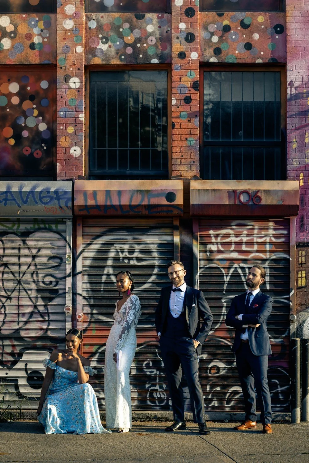 Four people in wedding attire posing against a brick facade and graffiti-covered shutters.