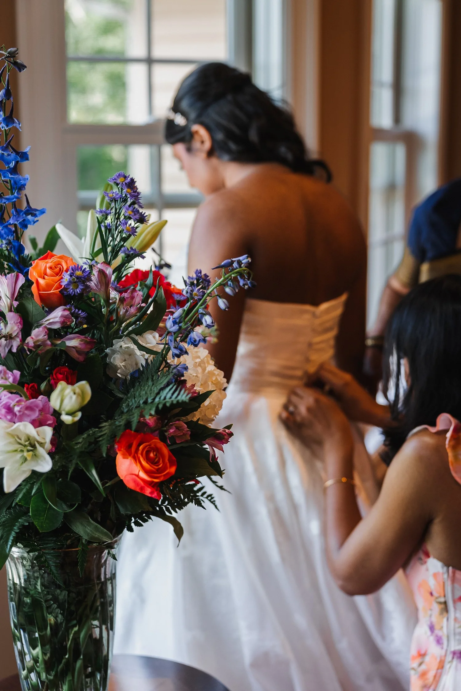 Natalia having the back of her wedding dress fastened beside a vase of bright flowers