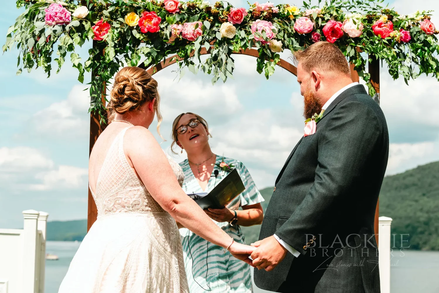 Amanda and Chris holding hands under the floral arbor during their ceremony at The Otesaga