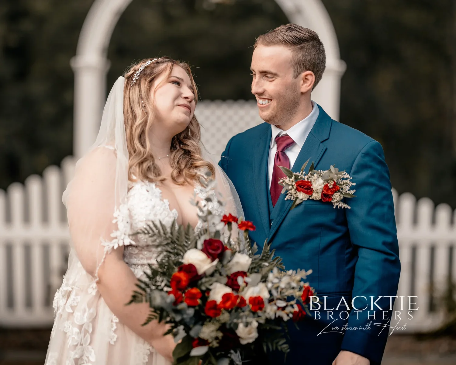 Jennifer and Jared standing close in front of the white arbor and picket fence, looking at each other and smiling, her bouquet between them