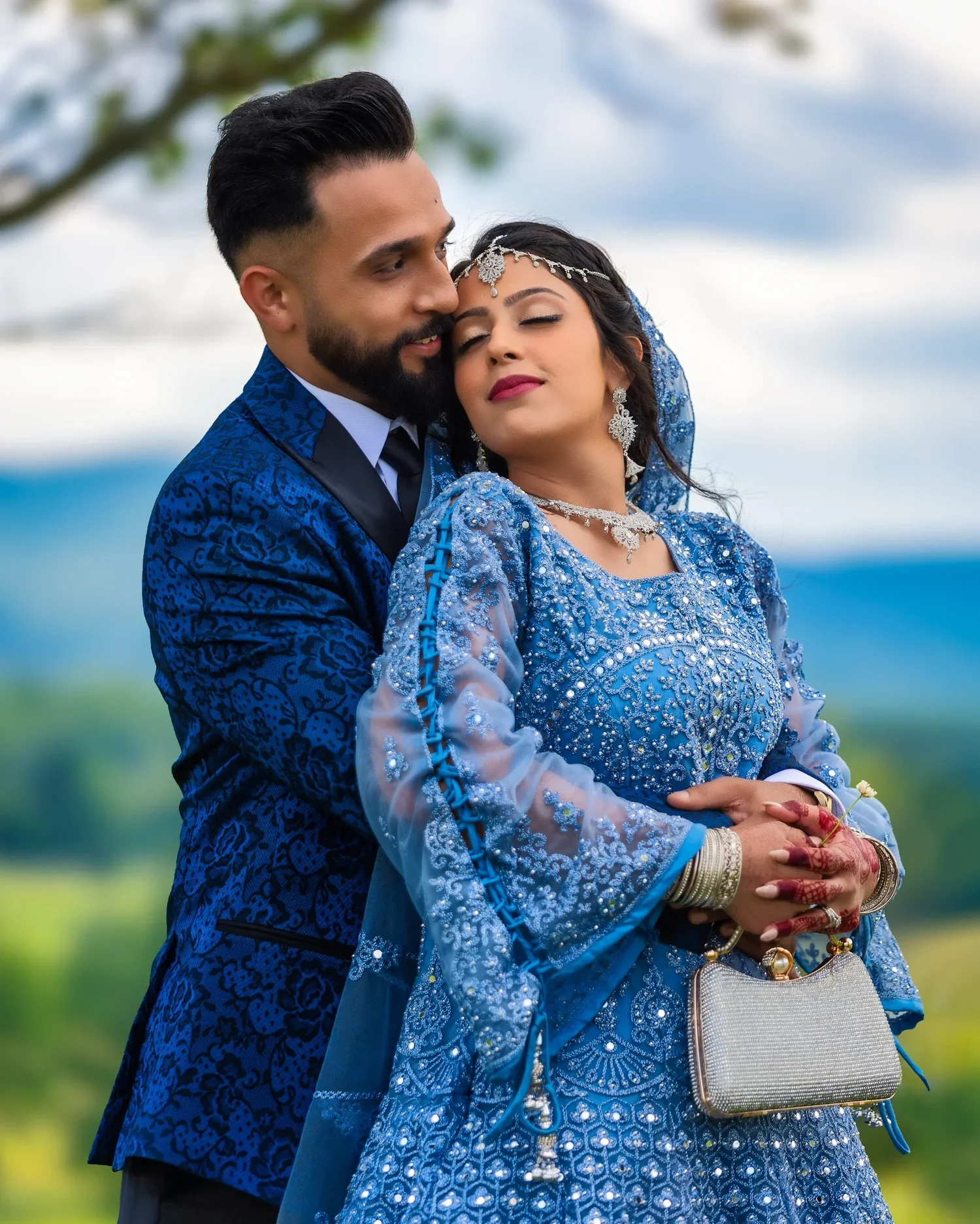 A couple standing close together on the hillside at Traditions at the Glen, the bride in a pale blue embroidered gown holding a silver clutch, hills soft in the distance