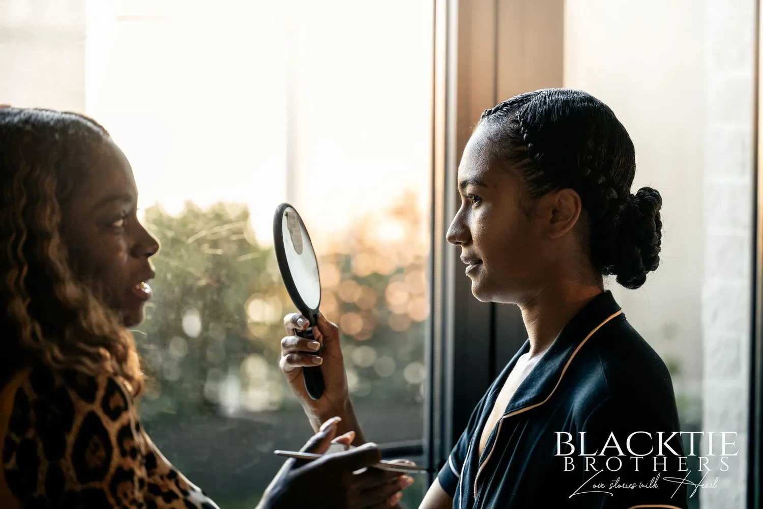 Ciara in profile holding a hand mirror by a bright window while her makeup artist looks on