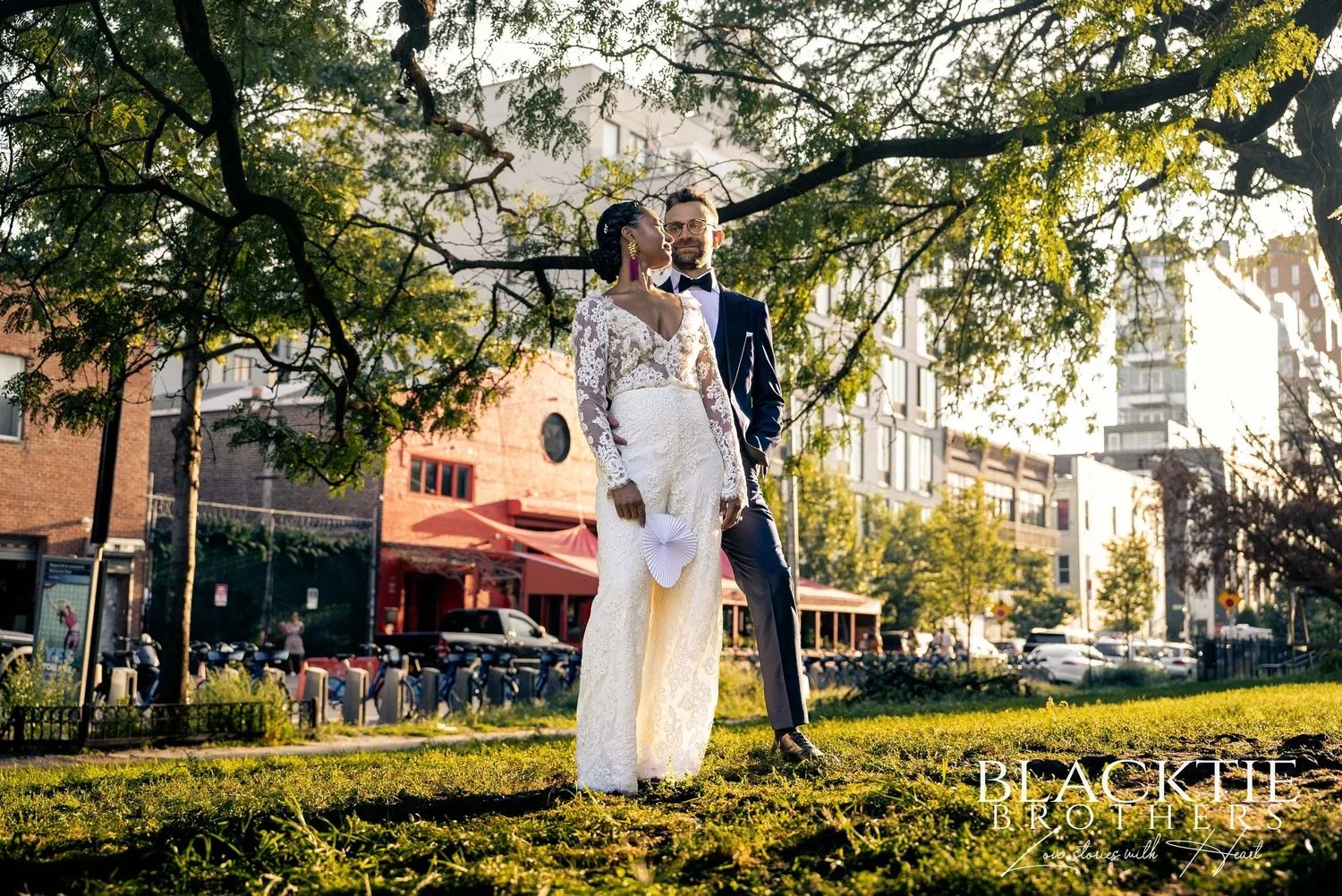Ciara and Drew standing on the grass in the park at golden hour under wide tree branches, red awnings and apartment buildings across the street