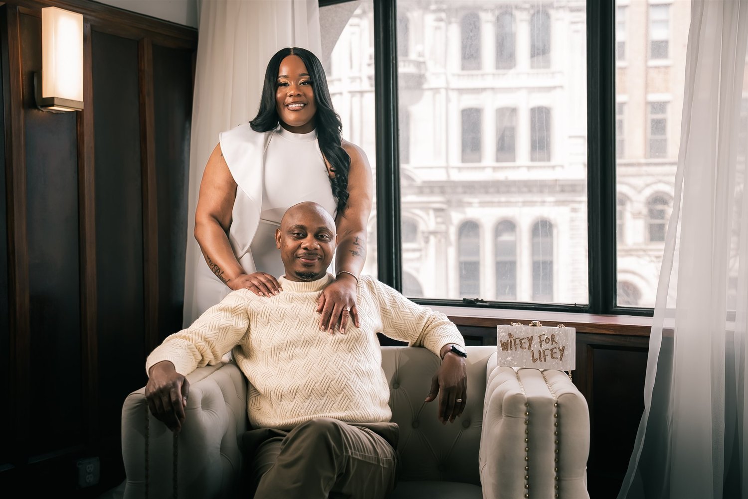 Tiffany stands behind David seated in an armchair beside a tall window during their second engagement session.