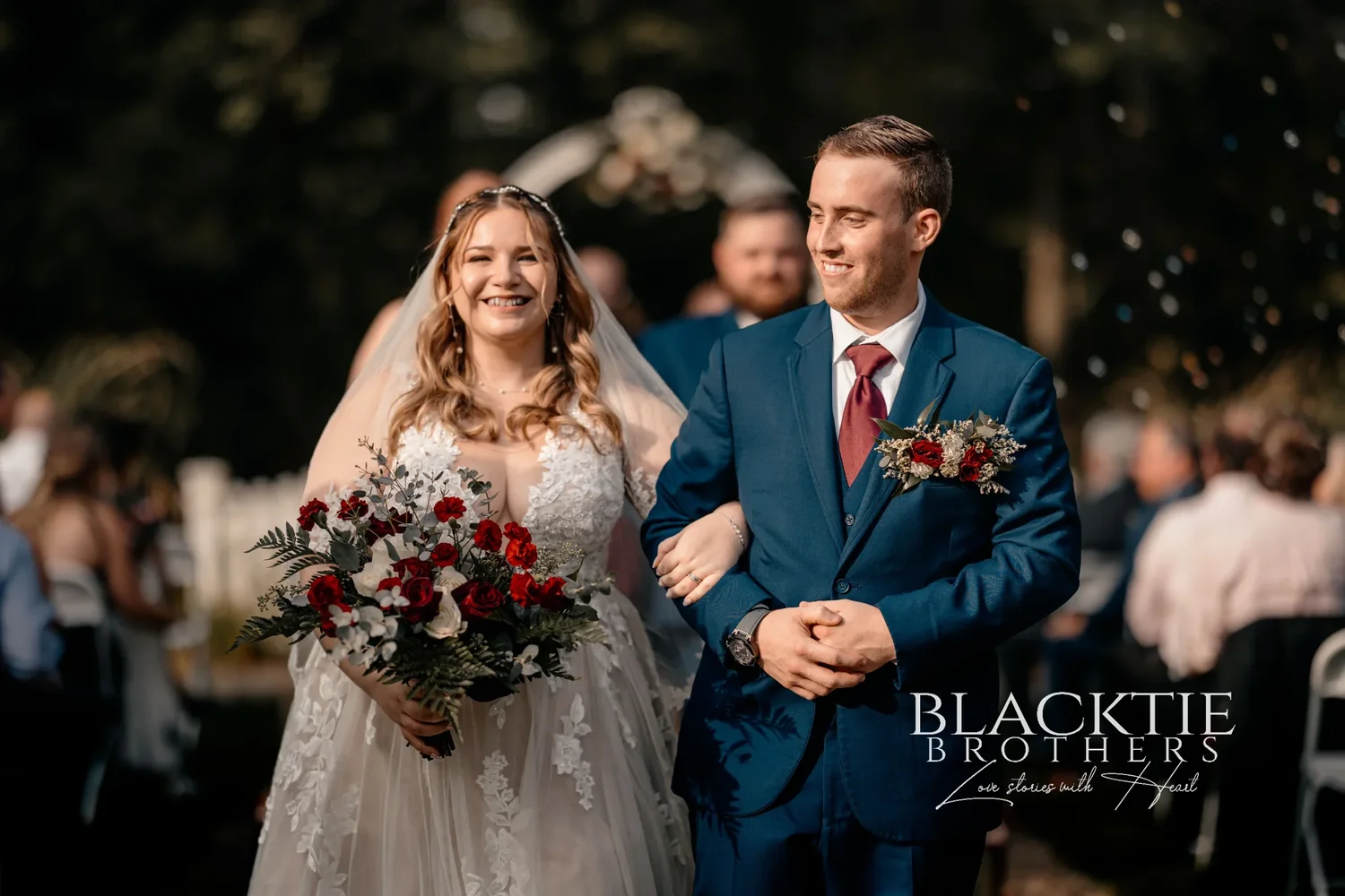 Jennifer laughing and Jared smiling as they walk arm in arm down the aisle after the ceremony, her red and white bouquet in front of them