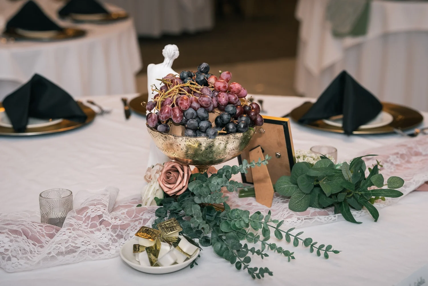 A centerpiece of grapes, roses and eucalyptus on a white tablecloth beside gold-rimmed plates