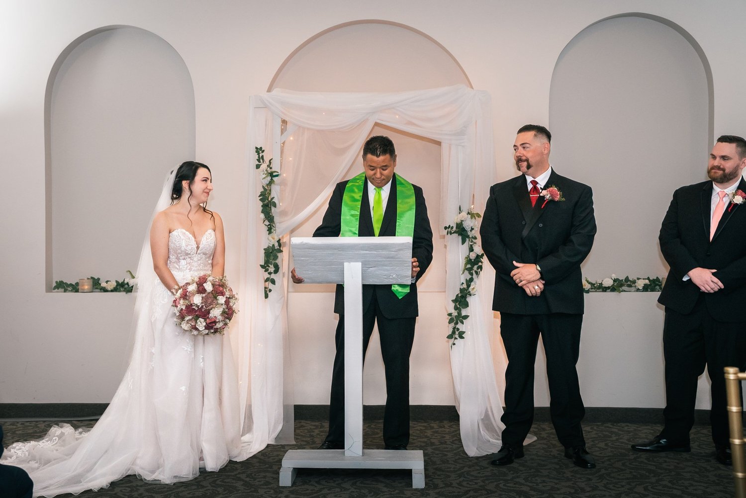 Laura and Dante facing their officiant at a white podium, framed by white arched alcoves and greenery
