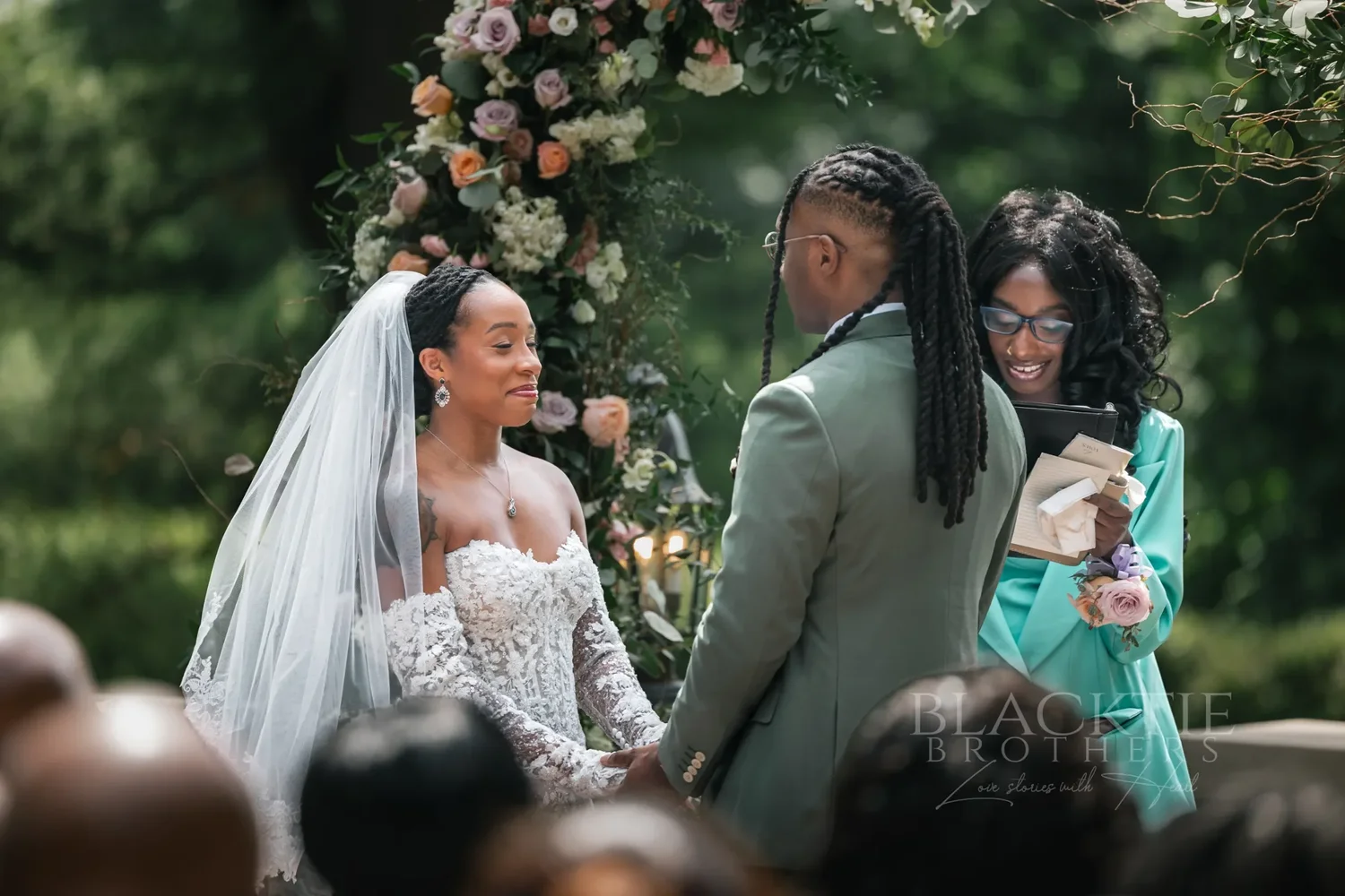 The bride and groom facing each other during the vows beside a floral arch, the officiant in a teal suit reading beside them