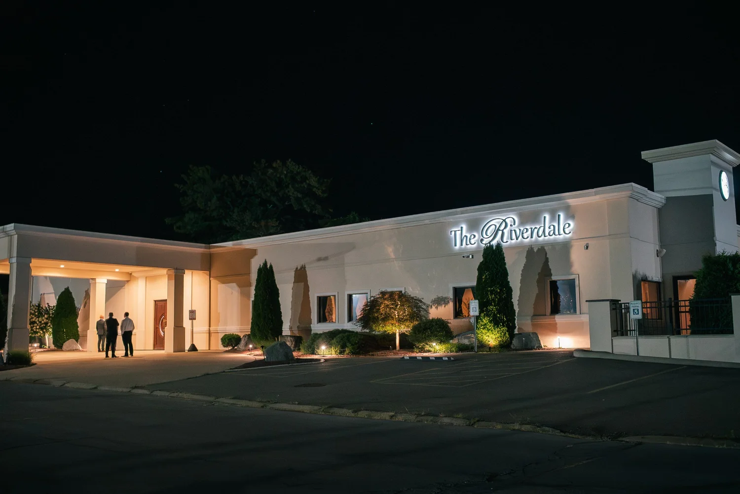 The Riverdale at night, its white facade and lit sign glowing above a quiet parking lot