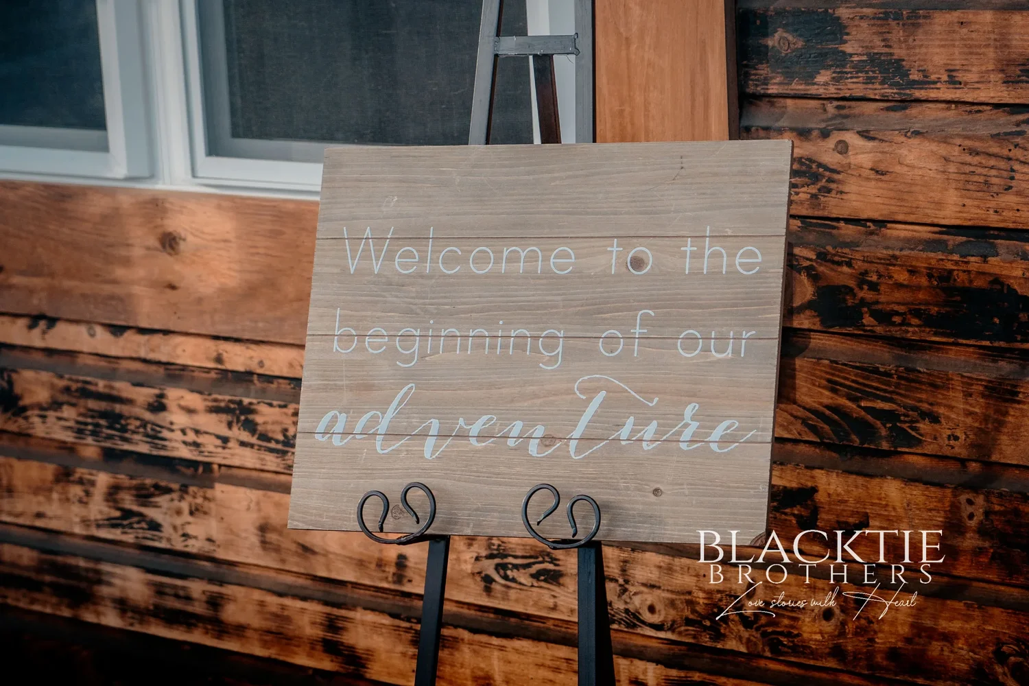 A wooden sign on an easel reading Welcome to the beginning of our adventure, against a dark timber wall