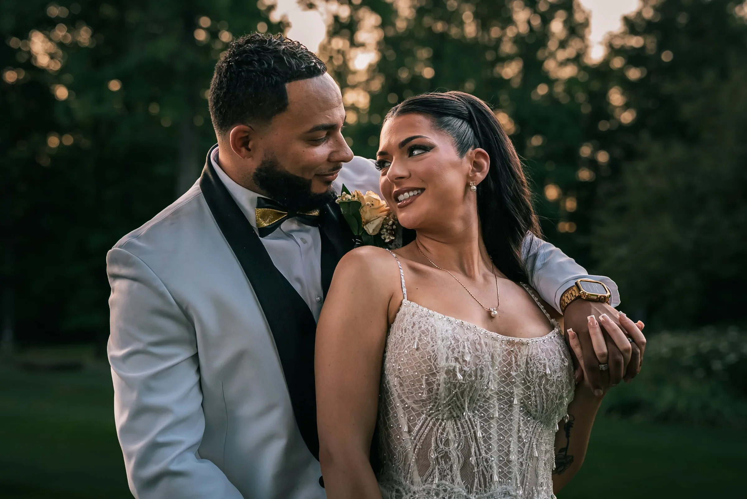 Trevor resting his arm on Vanessa’s shoulder as she looks back at him smiling, with golden light glowing through the trees