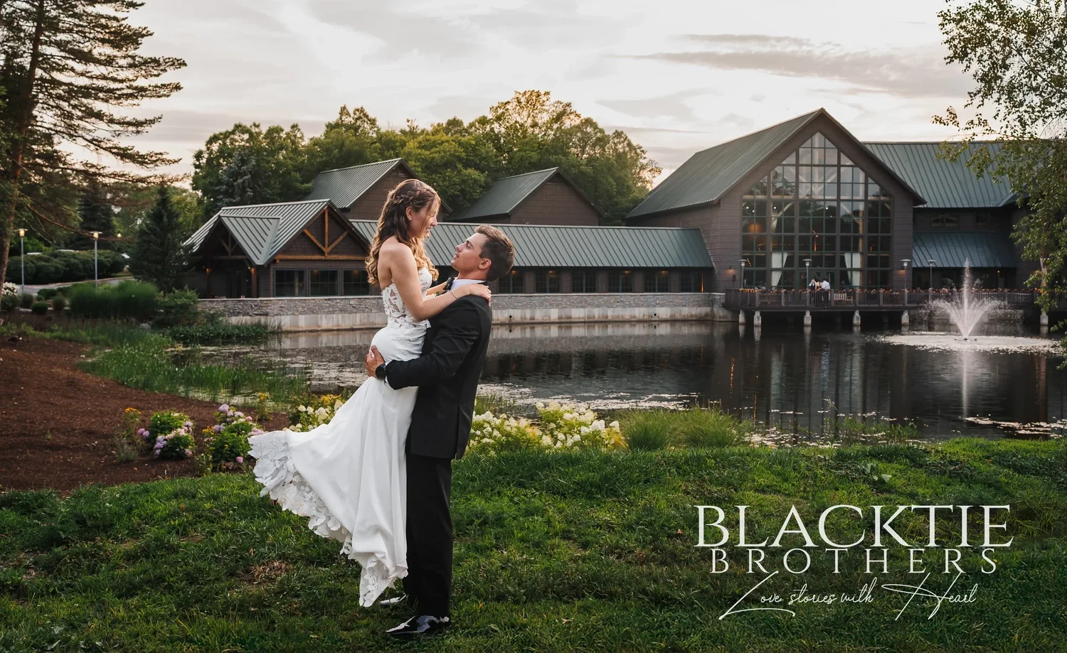 A groom lifting his bride on the far bank of the pond at Allyn Lodge, with the lodge and fountain behind them.