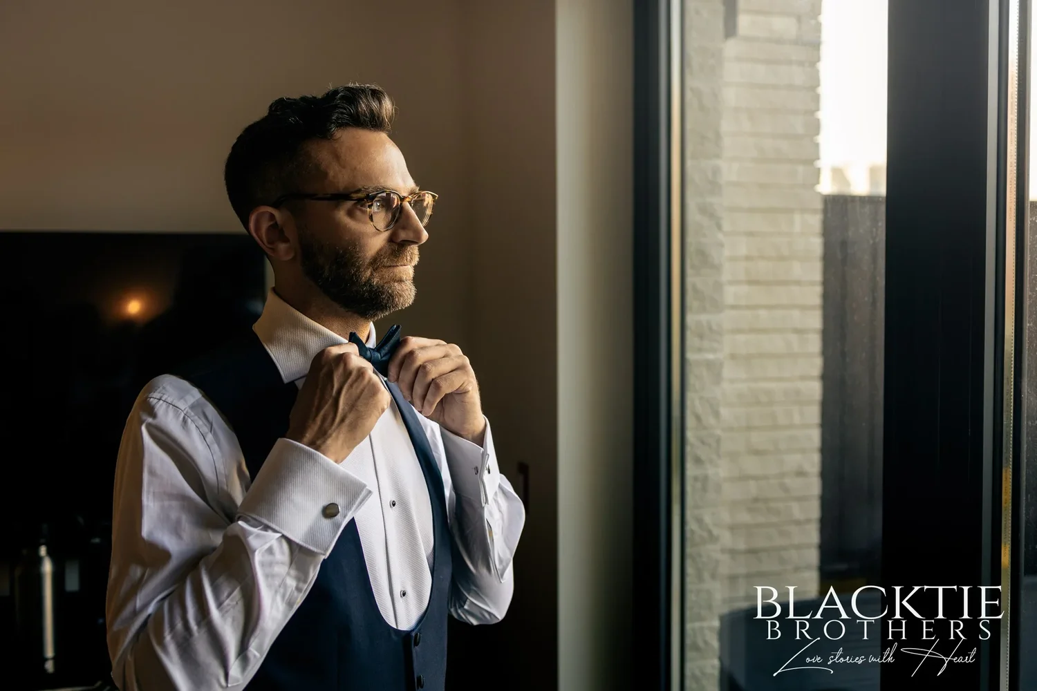 Drew in a white shirt and vest adjusting his bow tie beside a window
