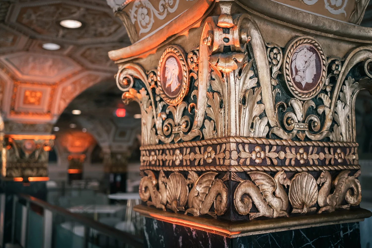 A gilded column capital with cameo medallions and carved sea creatures inside Palladian Hall, lit in warm orange.
