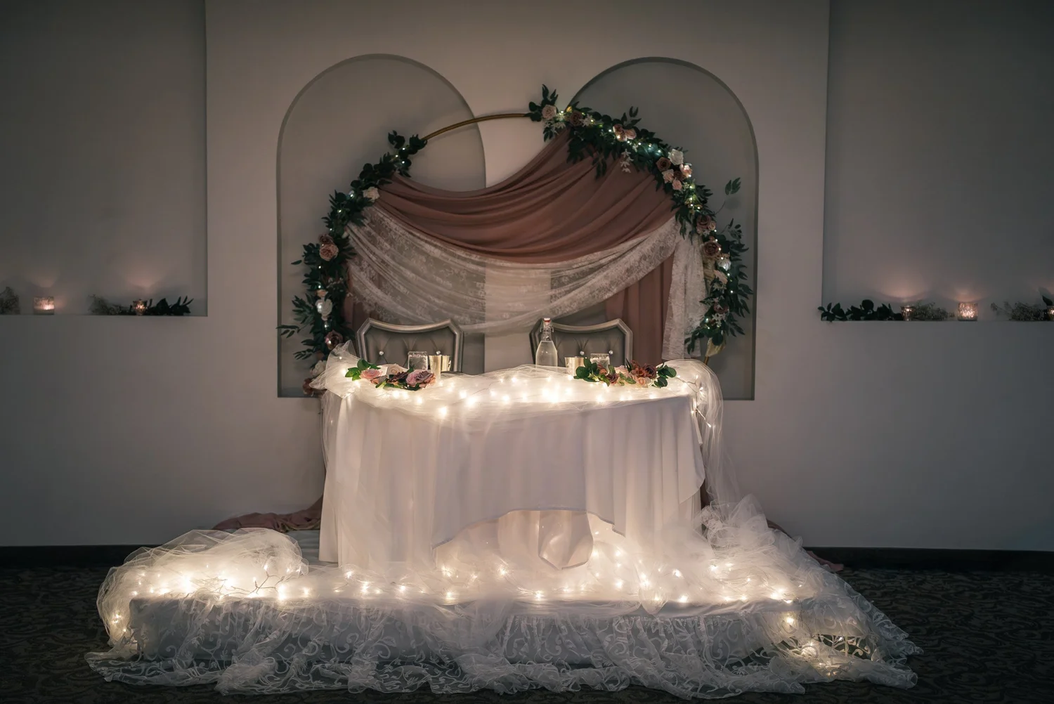 The sweetheart table under a pink and white draped arch wrapped in roses and greenery, string lights glowing along the front
