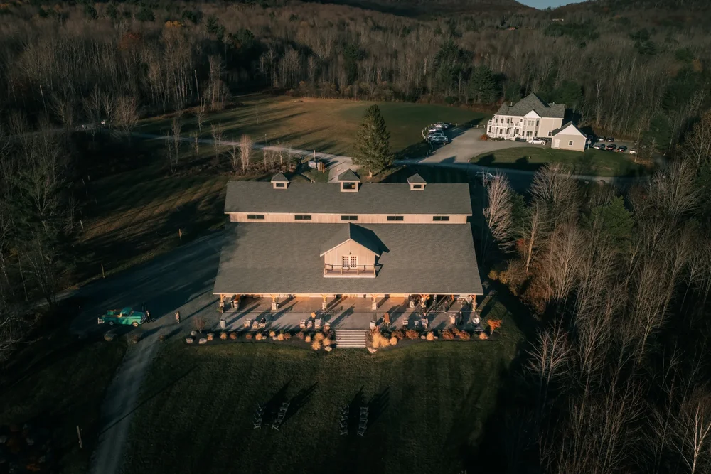 Windham Manor from above in autumn: the timber Barn and its covered patio in the foreground, the white Victorian Manor House beyond it and the wooded Catskill Mountains all around