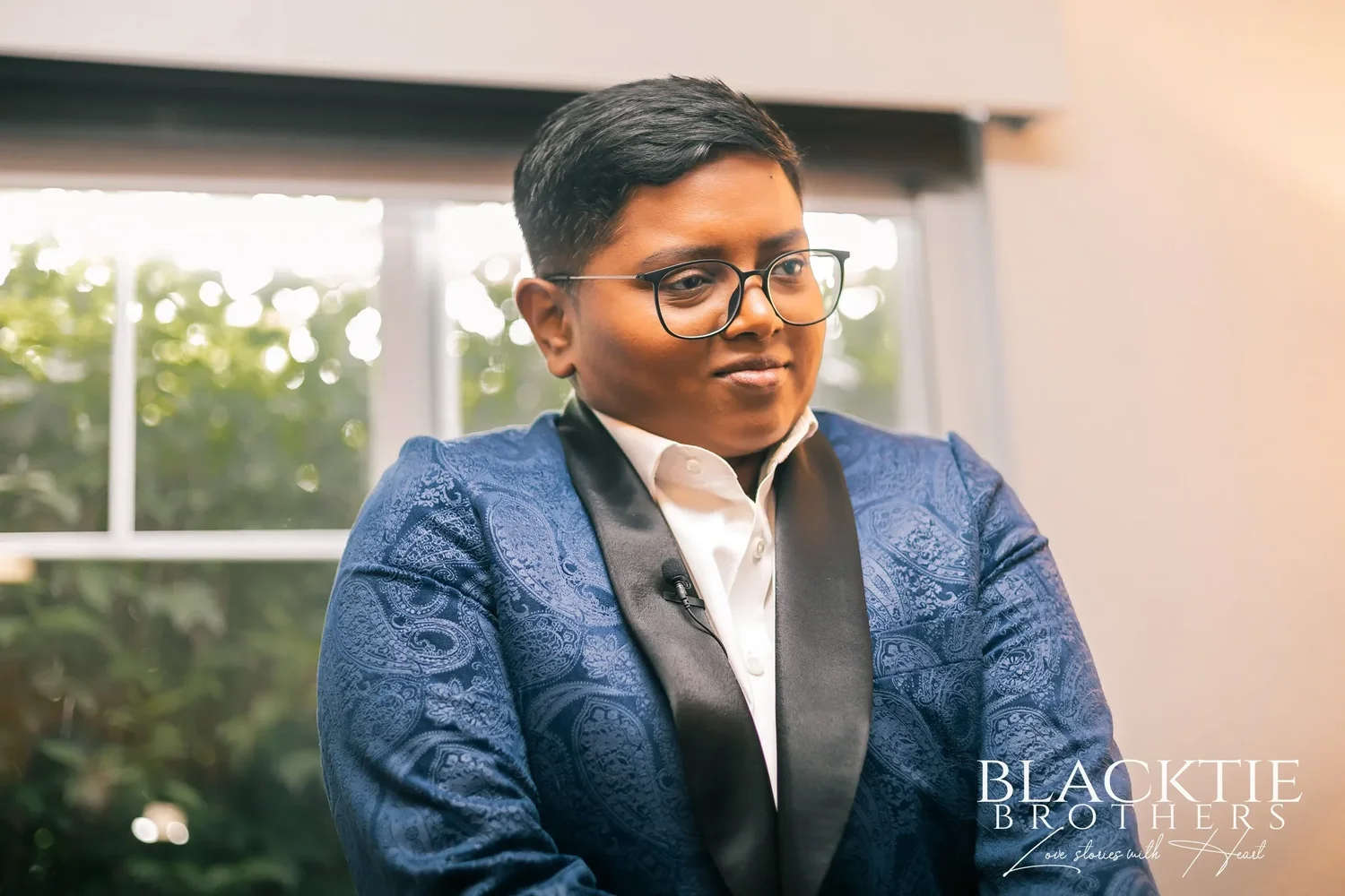 Portrait by a window of one of the couple in a blue paisley tuxedo jacket with black lapels and glasses, smiling slightly