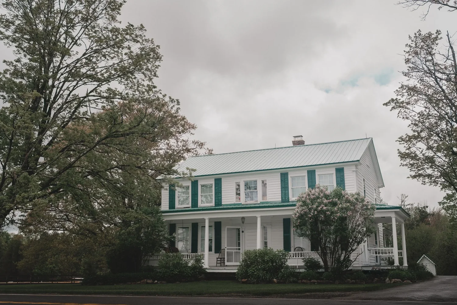 The white farmhouse at Knotting Hill Farm with green shutters, a wraparound porch and tall trees