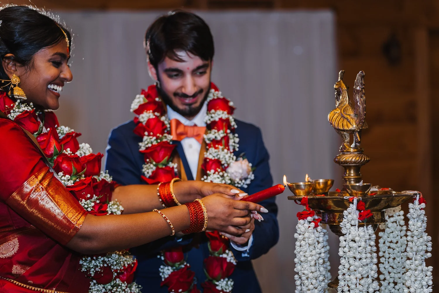 Natalia and Ansel, both wearing flower garlands, lighting a tall brass lamp together