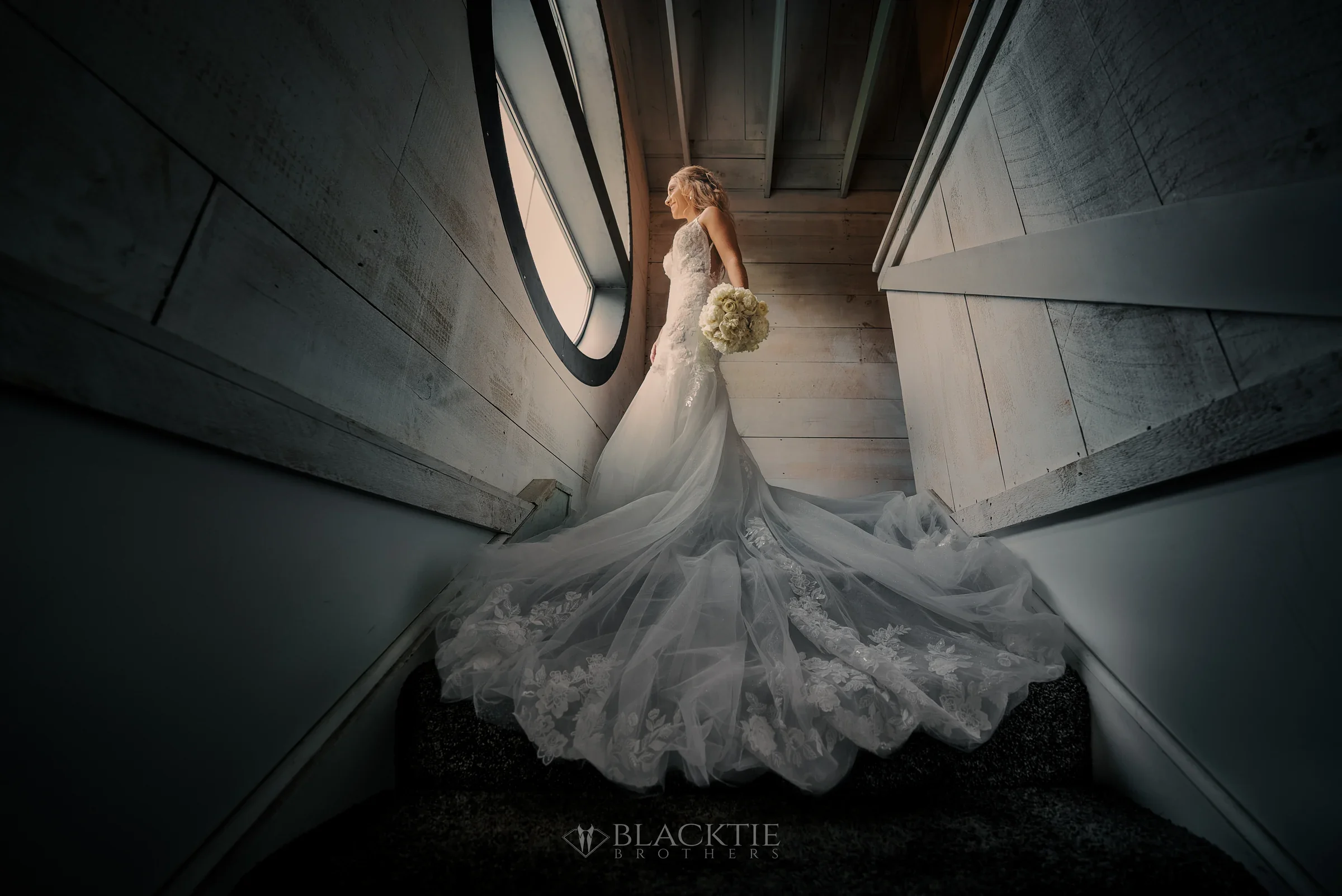 Jennifer in her wedding dress at the top of a whitewashed wood stairwell at The Cannery, lit from a round window beside her, her lace train spread down the steps and a white bouquet in her hand