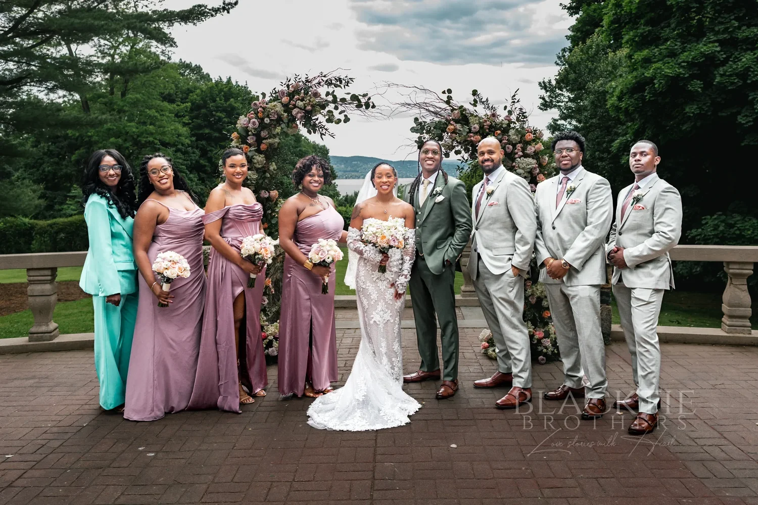 The couple with their wedding party lined up along the stone balustrade between the floral arches, bridesmaids in mauve and groomsmen in light gray suits