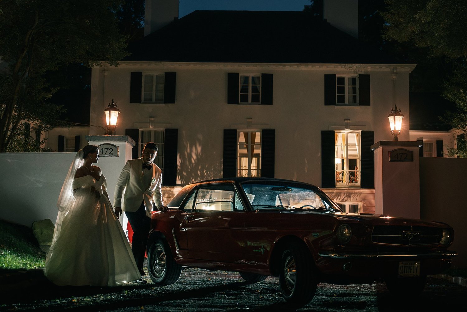 Patricia and Joshua beside a red vintage Mustang in front of a lit white house at night.