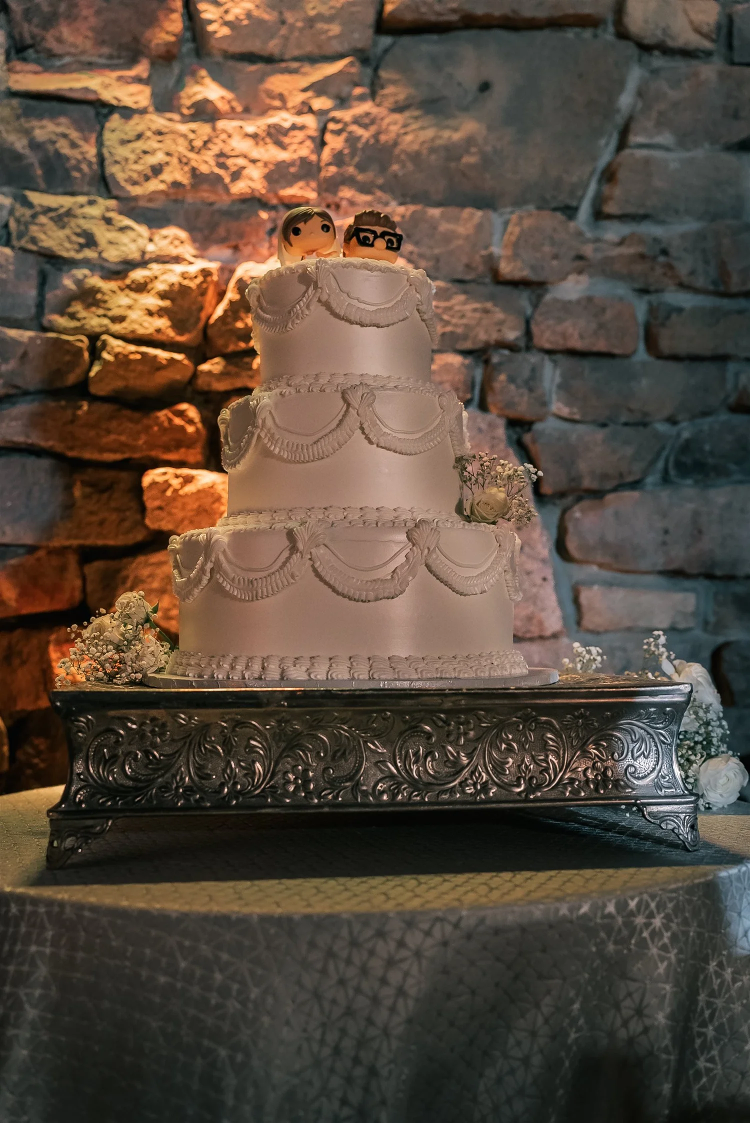 A tiered white wedding cake on a silver stand against the stone wall at The Somerley.