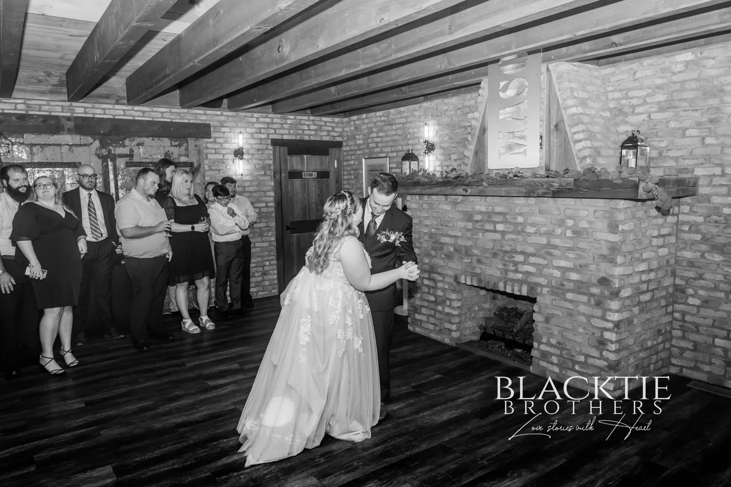 Black and white view of the reception room at The Mason Jar: exposed brick, timber ceiling beams, a brick fireplace and guests watching the couple dance