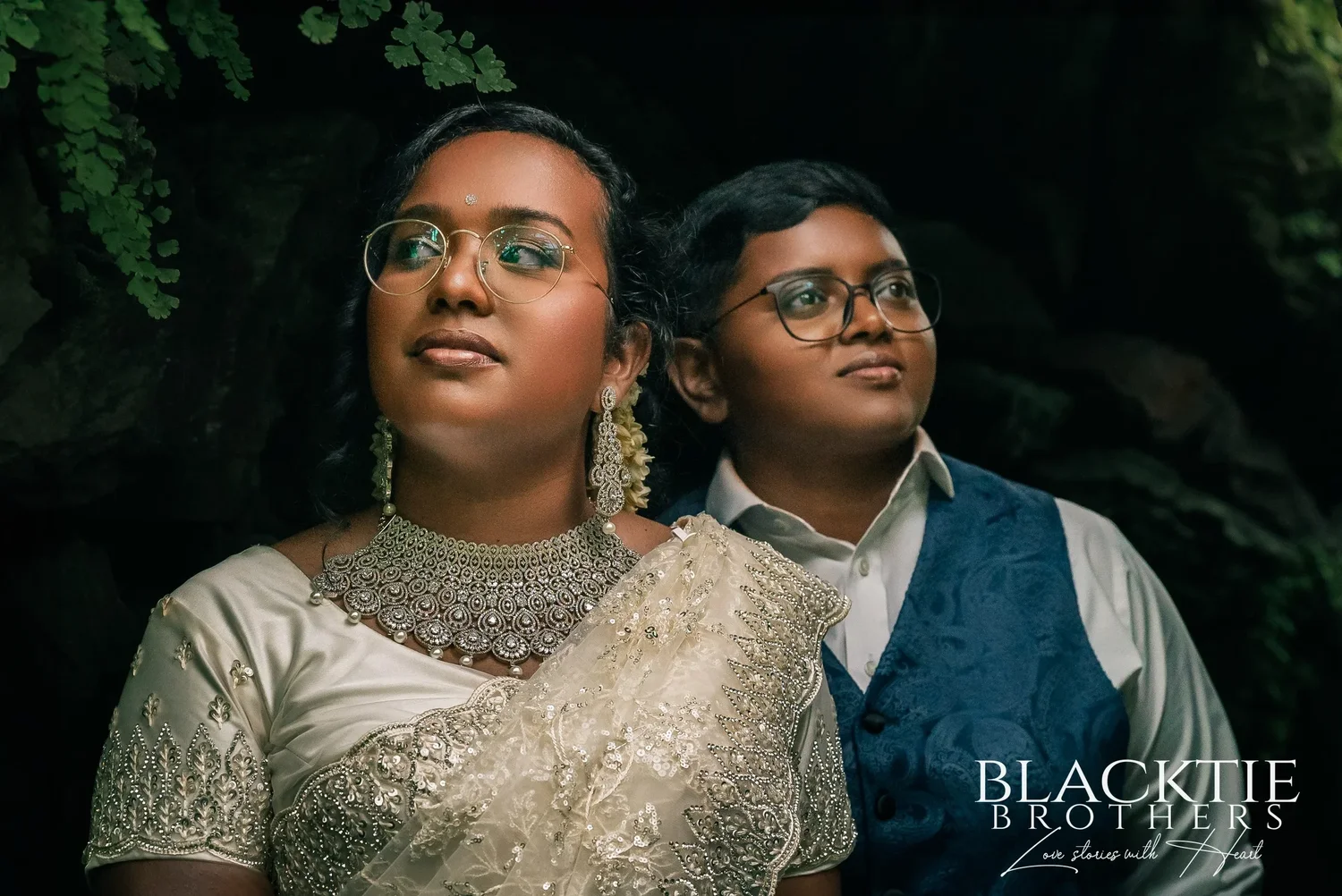 Hima and Alina looking up and to the side under an overhang of ferns, one in a silver choker and long earrings, one in a blue paisley vest