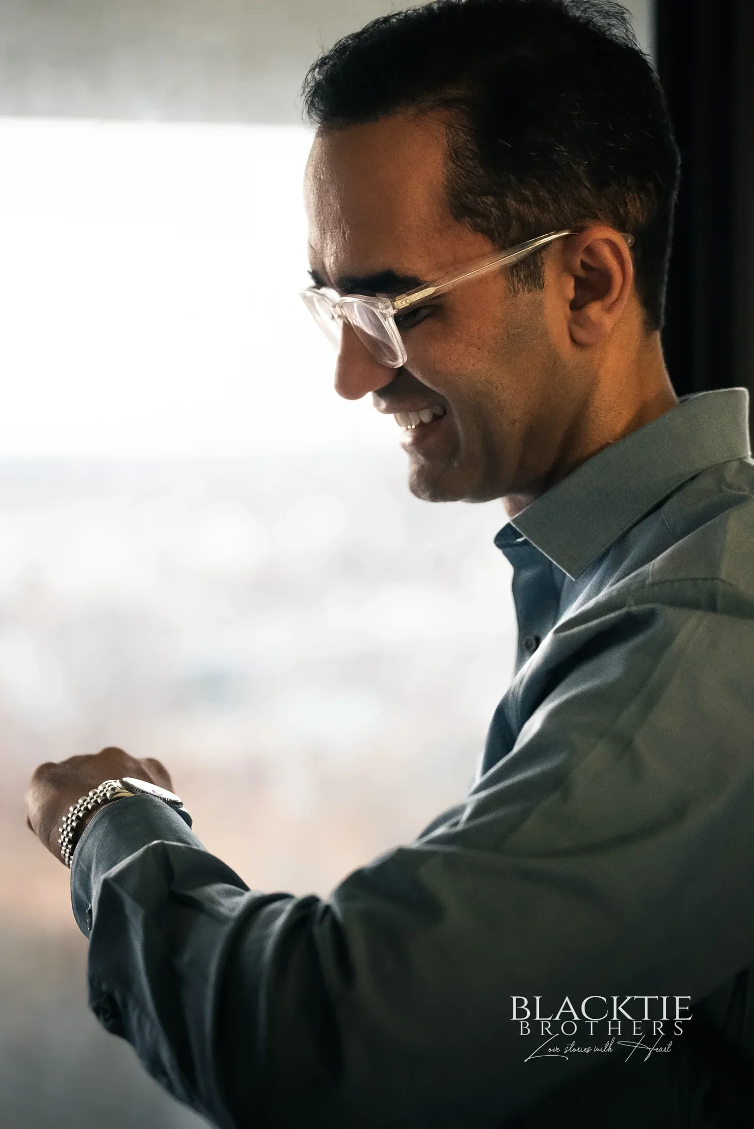 Rizwan in a blue shirt and glasses fastening his watch beside a bright hotel room window, smiling down at his wrist